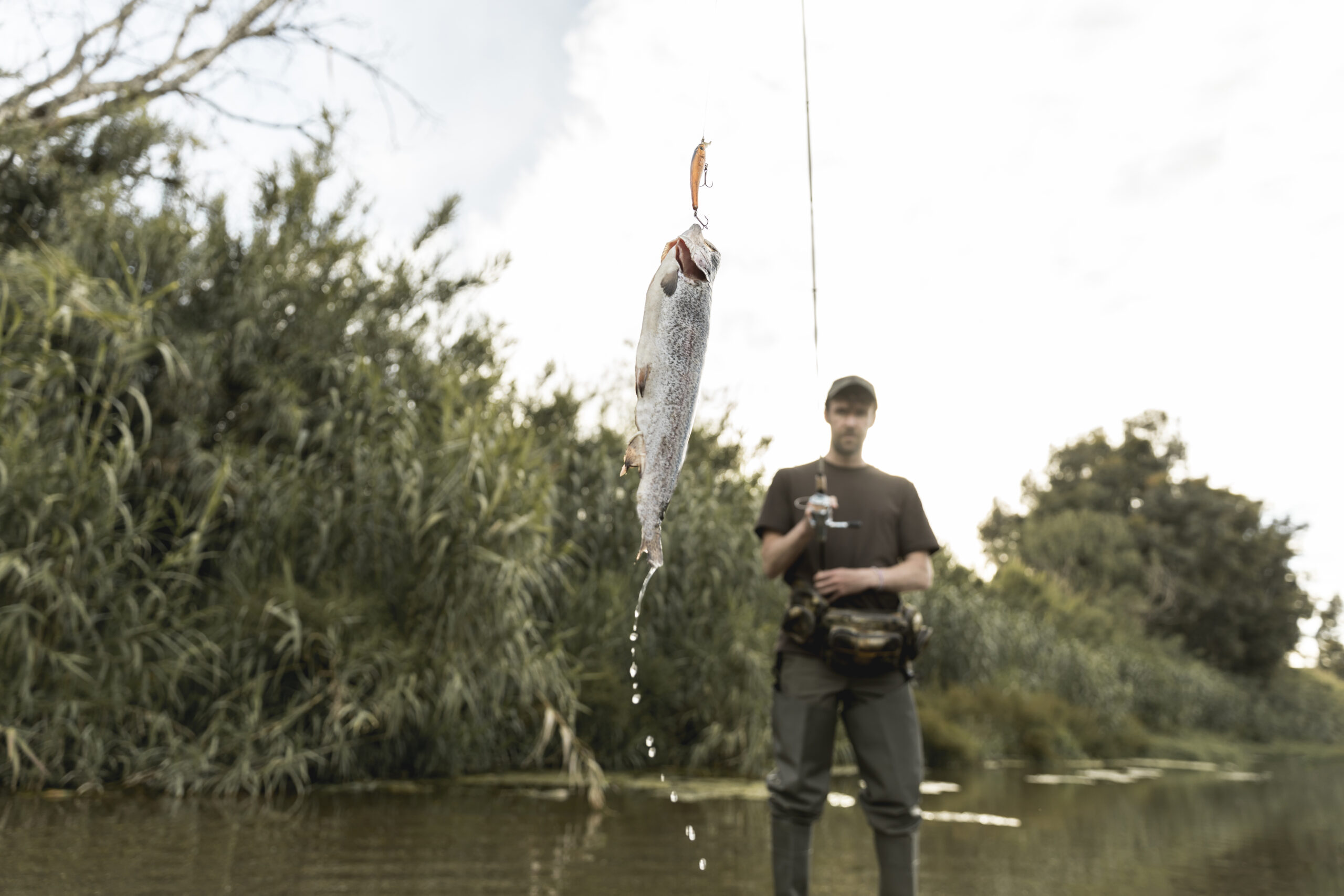 Angler in waders holding a fly fishing rod and reel with a freshly caught fish hanging from the line by a river